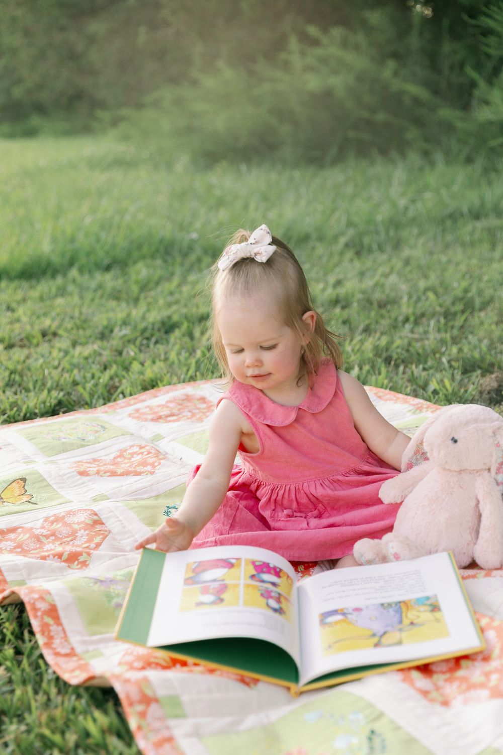 Adorable toddler girl in pink dress reads picture book on quilt in green grass with stuffed bunny.