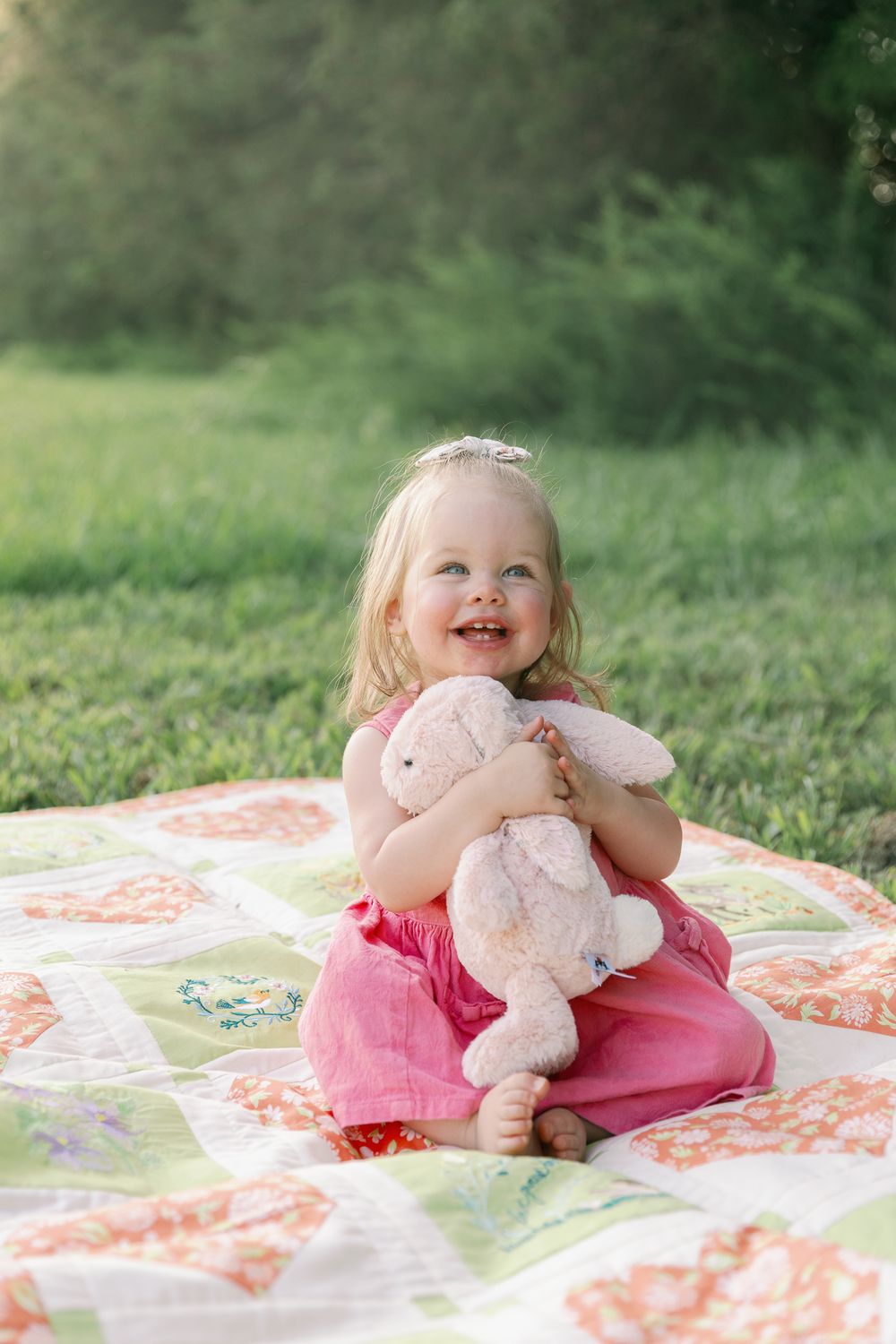Happy toddler girl in pink dress hugging stuffed bunny on colorful quilt in sunny green garden.