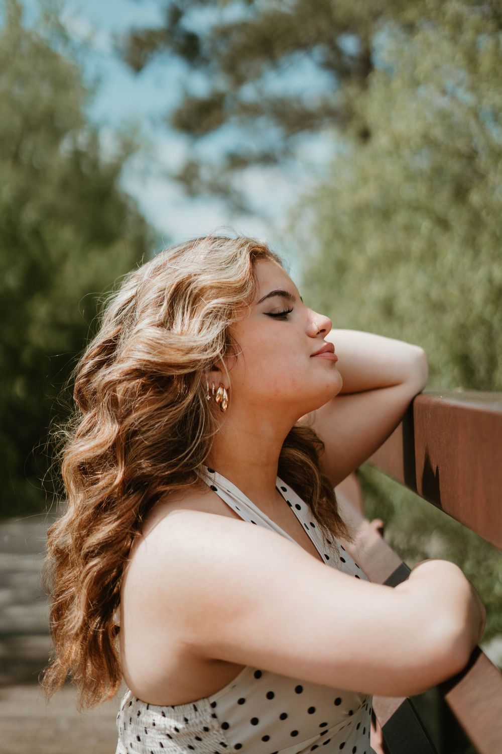 Young woman with curly blonde hair relaxing outdoors in polka dot top, eyes closed, leaning on wooden railing.