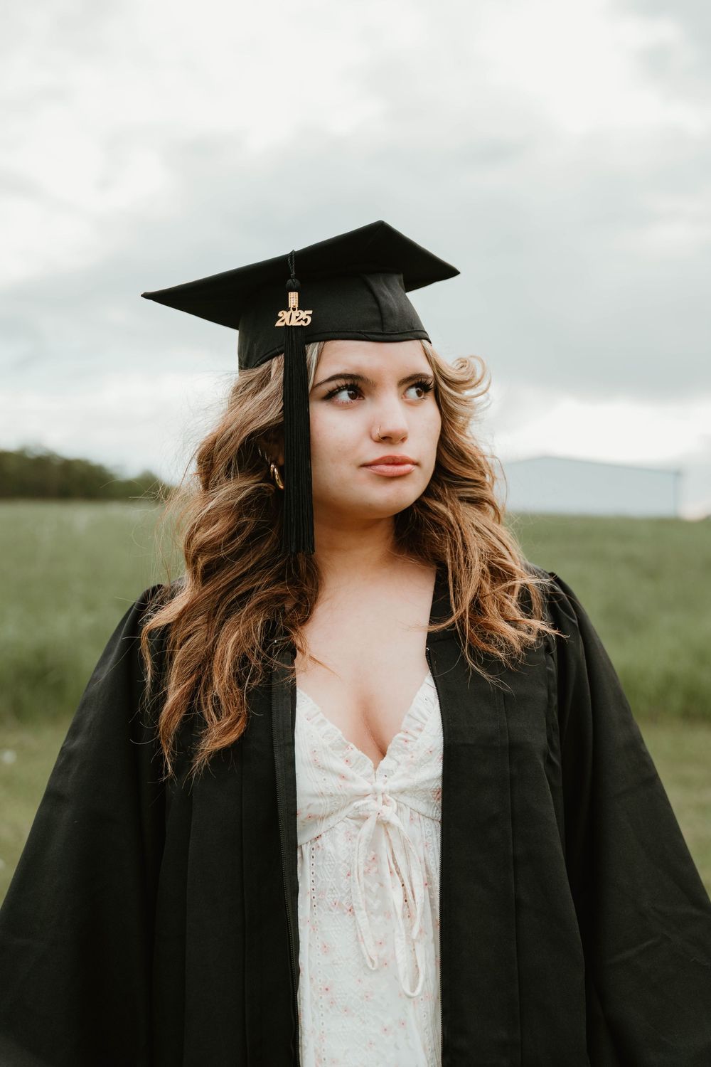 Young woman in black graduation cap and gown for class of 2025, posing outdoors with cloudy sky background.