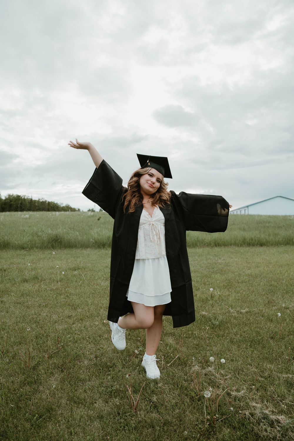 Young woman in black graduation gown and cap posing joyfully in a green field on a cloudy day.