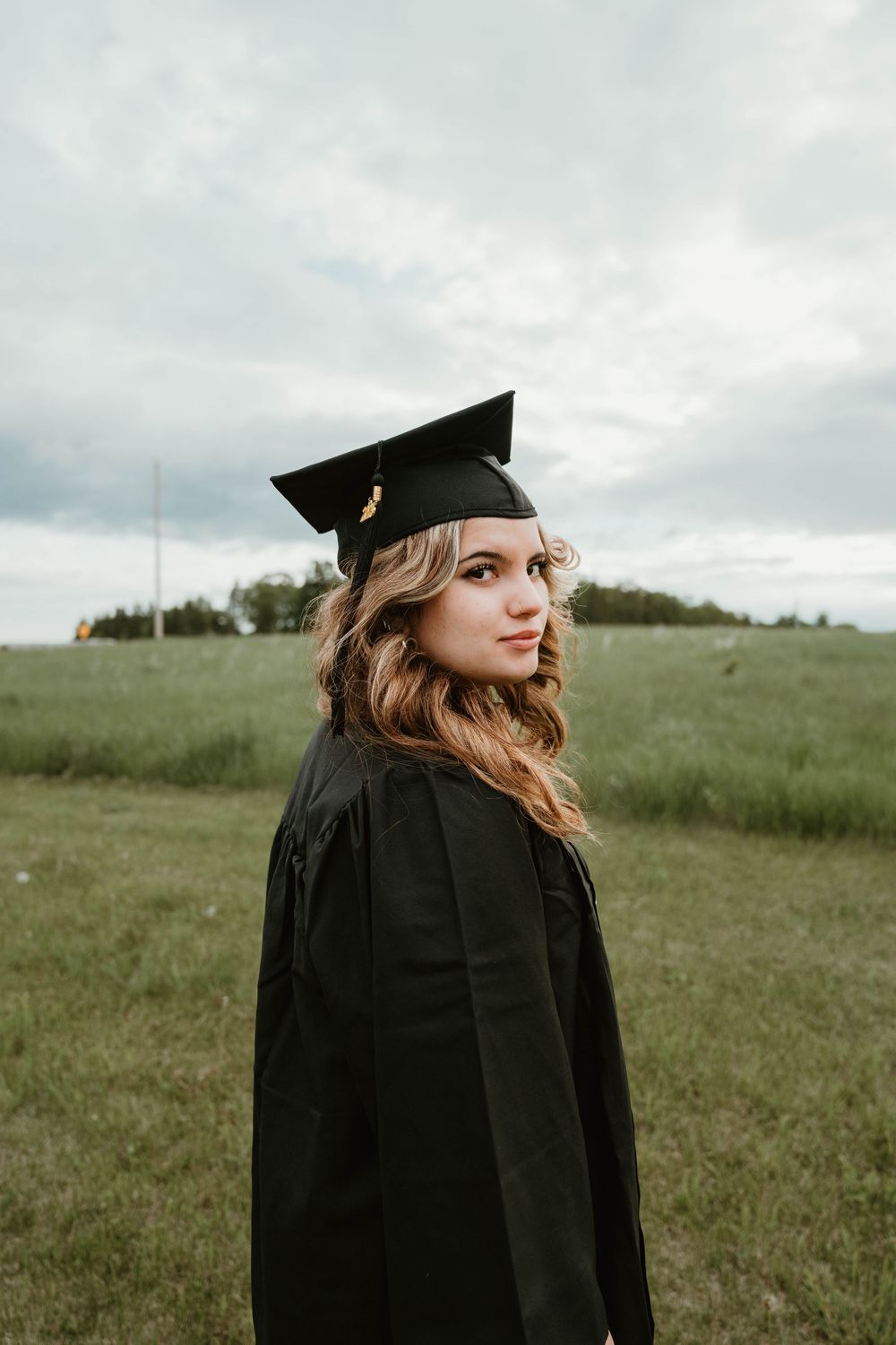 Young woman in black graduation cap and gown smiling outdoors in a green field on a cloudy day.
