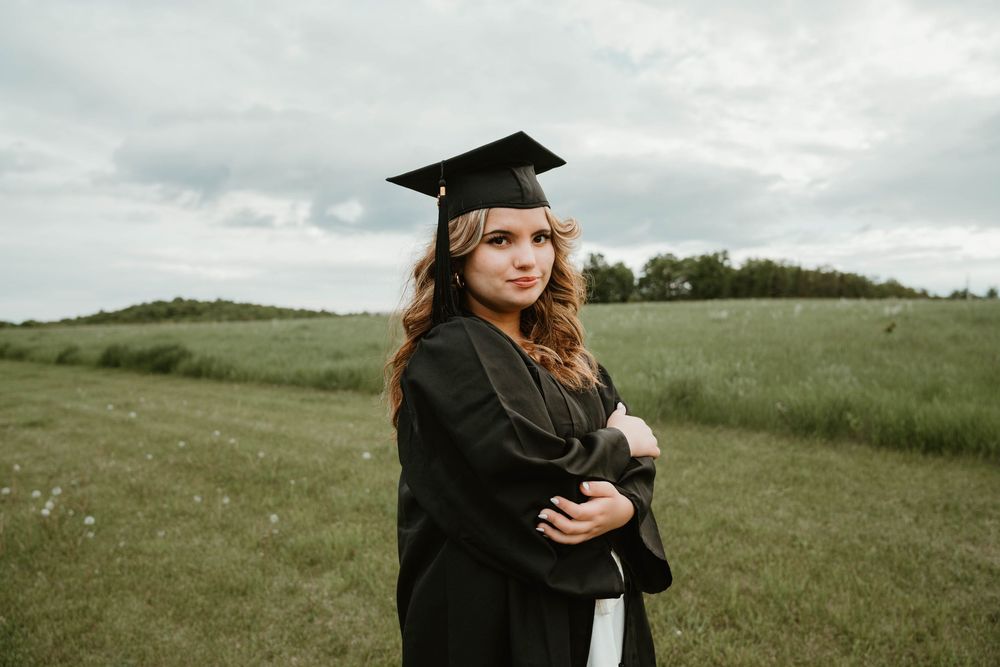 Young woman in black graduation cap and gown standing in green field, holding diploma, cloudy sky background.