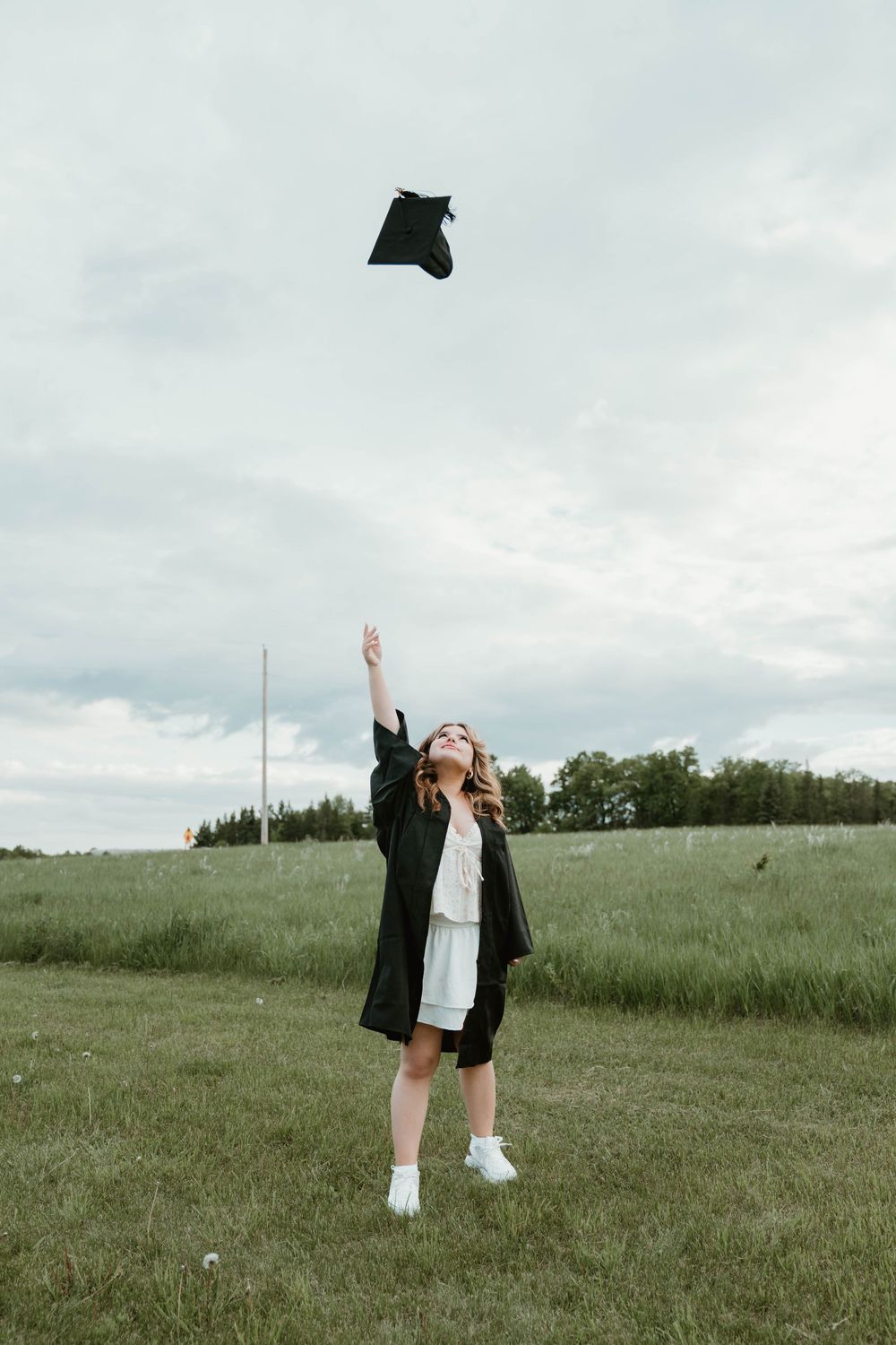 A graduate in black gown tosses her cap in the air outdoors in a green field under a cloudy sky.