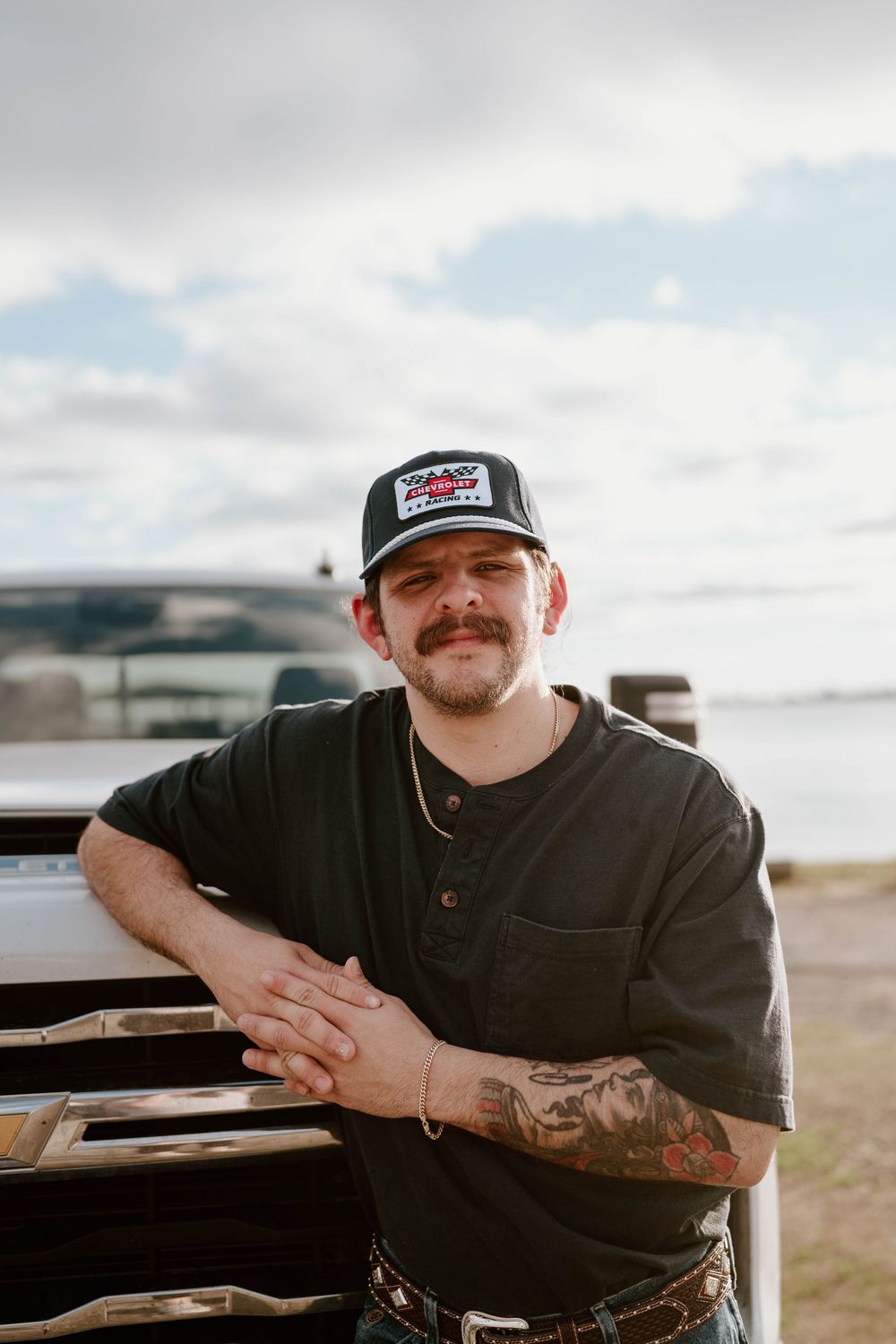 Tattooed man with mustache leaning on truck hood wearing black henley and trucker cap outdoors.