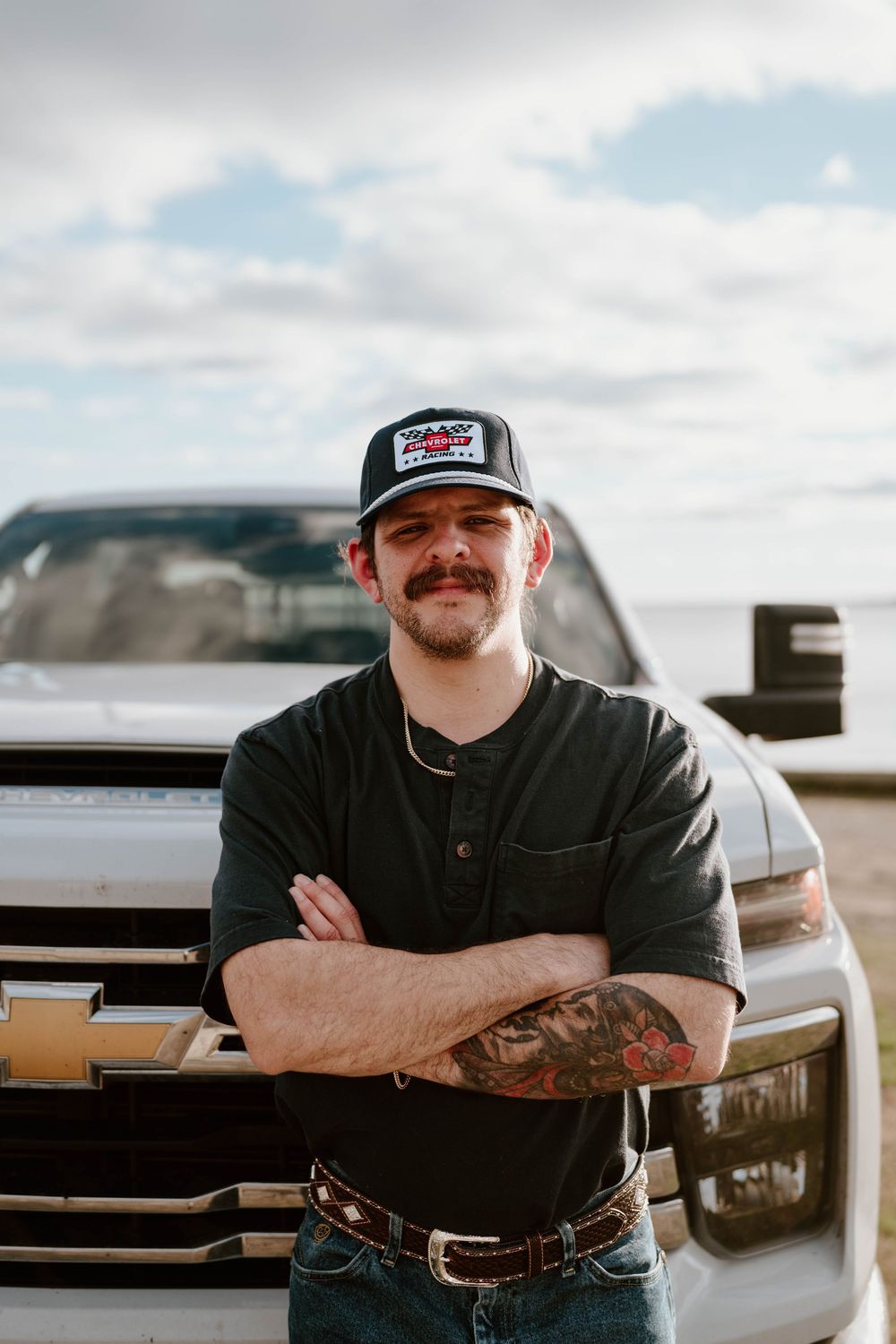 Man with mustache, tattoos, and trucker hat stands arms crossed in front of Chevrolet truck outdoors.