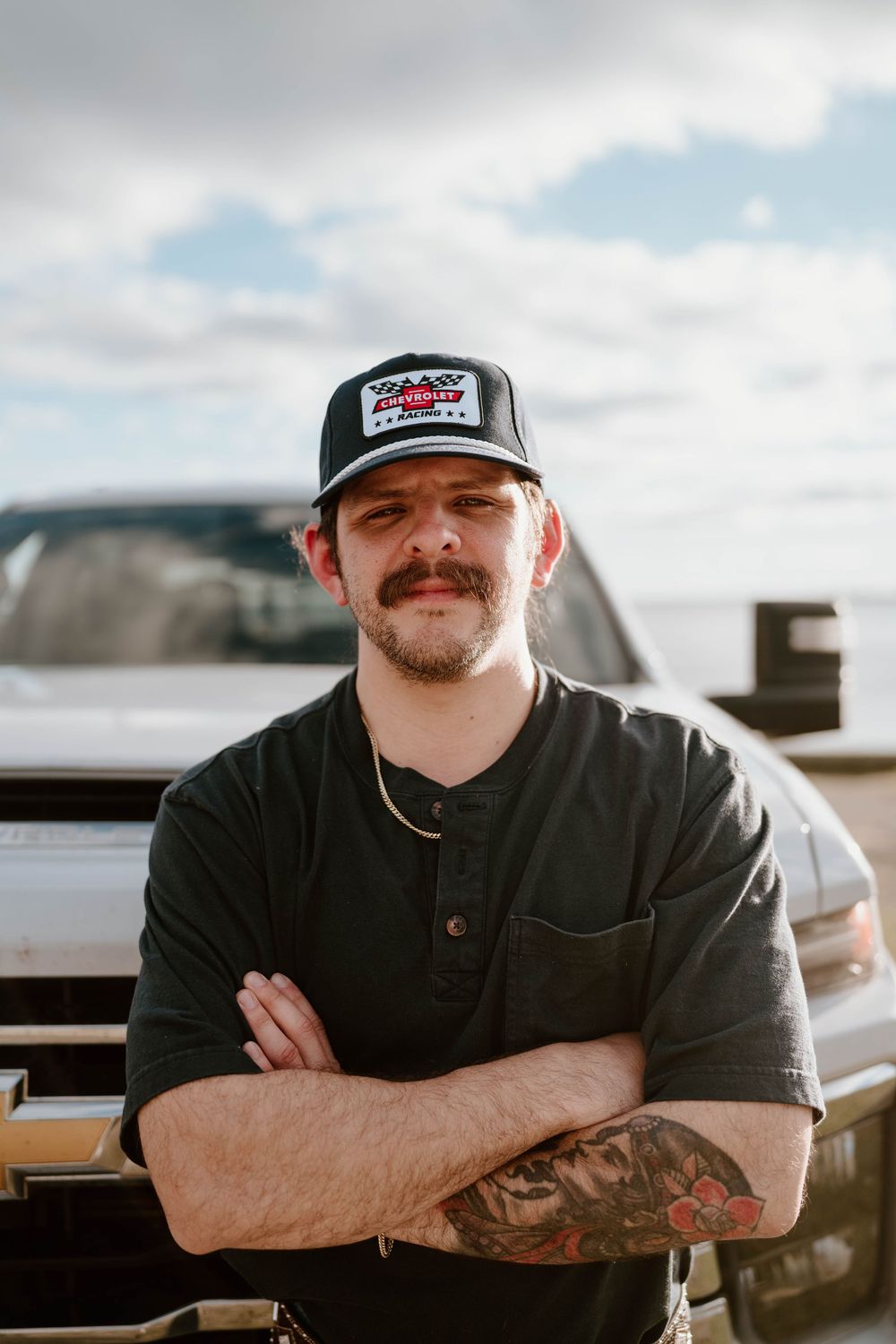 Tattooed man with mustache, crossed arms, wearing black cap and shirt, standing in front of a Chevrolet truck outdoors.