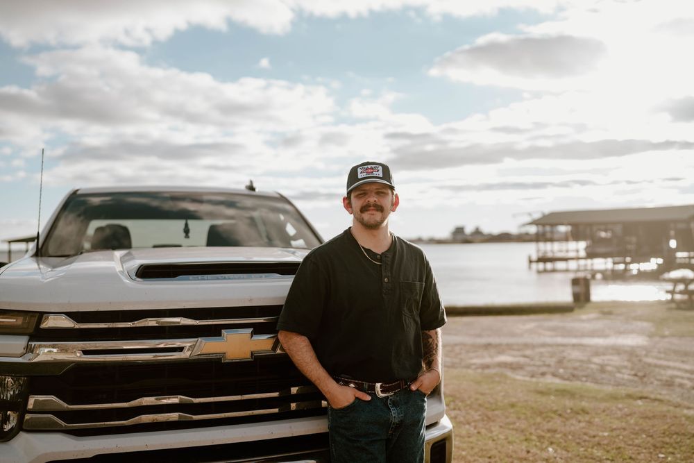 Man in black shirt and cap standing in front of Chevrolet truck near waterfront on cloudy day.