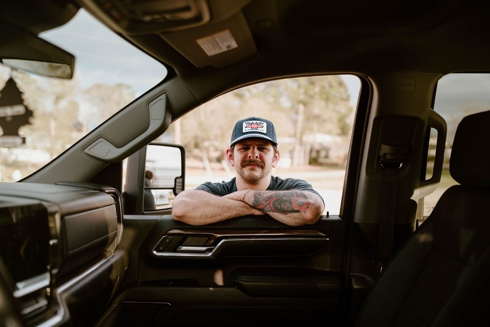 Man in trucker hat leans on truck window with arms crossed, showing tattoos, smiling casually.