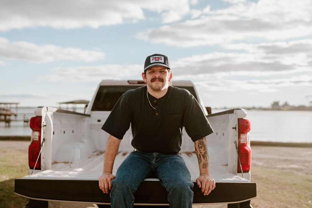 Man with mustache sits on white pickup truck bed near waterfront wearing black tee and jeans.