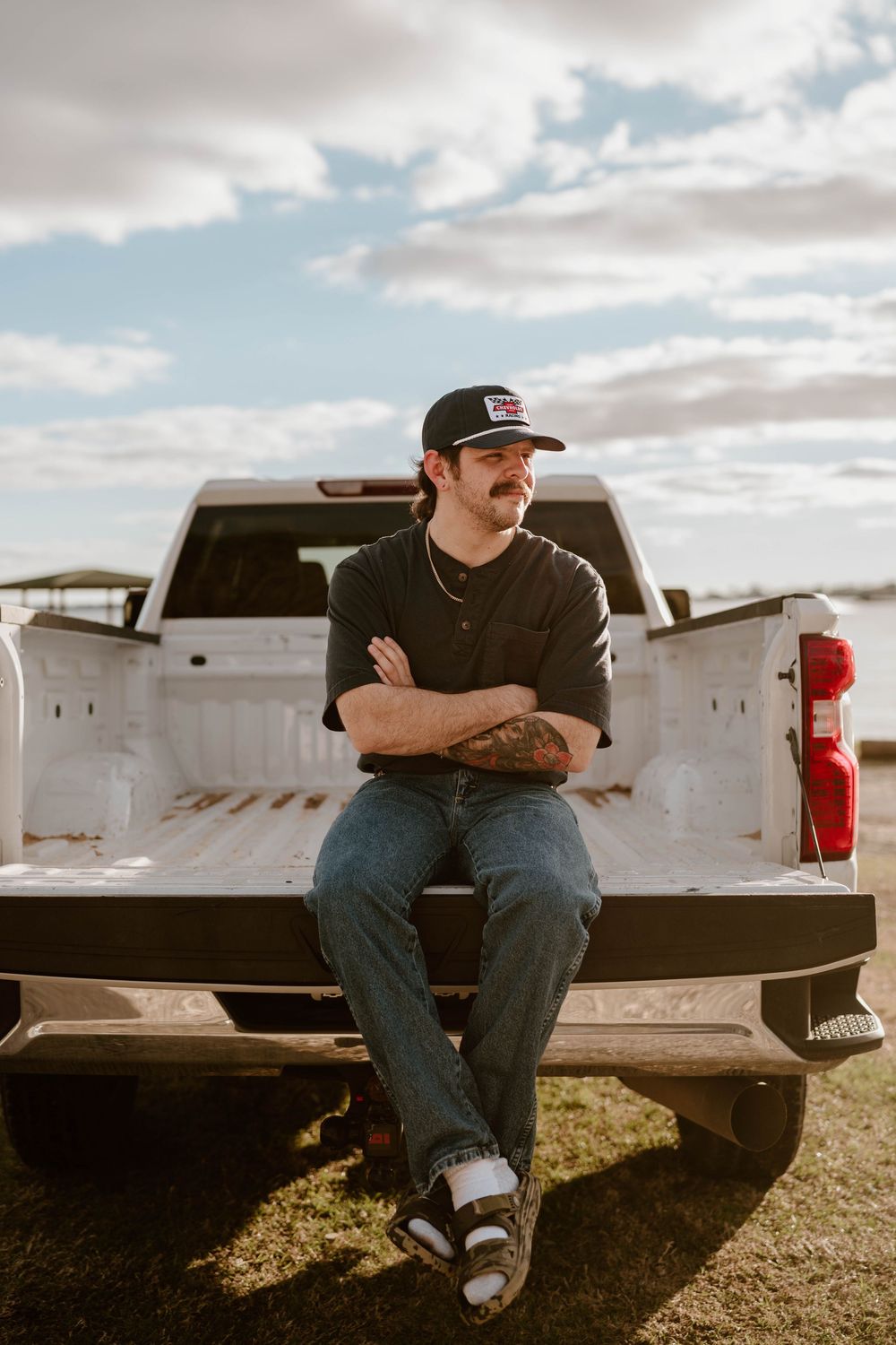Man with tattoos sitting on white pickup truck tailgate, wearing black cap and jeans, arms crossed outdoors.