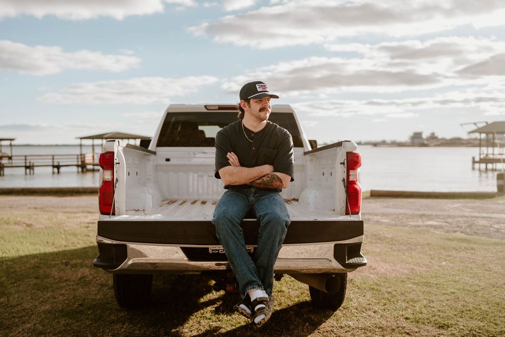 Man sitting on tailgate of pickup truck near waterfront, wearing cap and casual clothes on cloudy day.