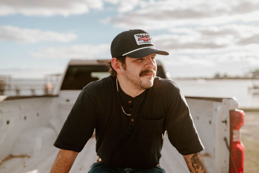 Tattooed man in black cap and shirt leans on truck bed, gazing sideways against cloudy sky.