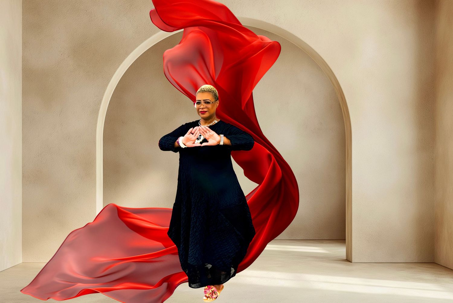 Woman in black dress poses gracefully with flowing red fabric billowing around her against a neutral arched backdrop.