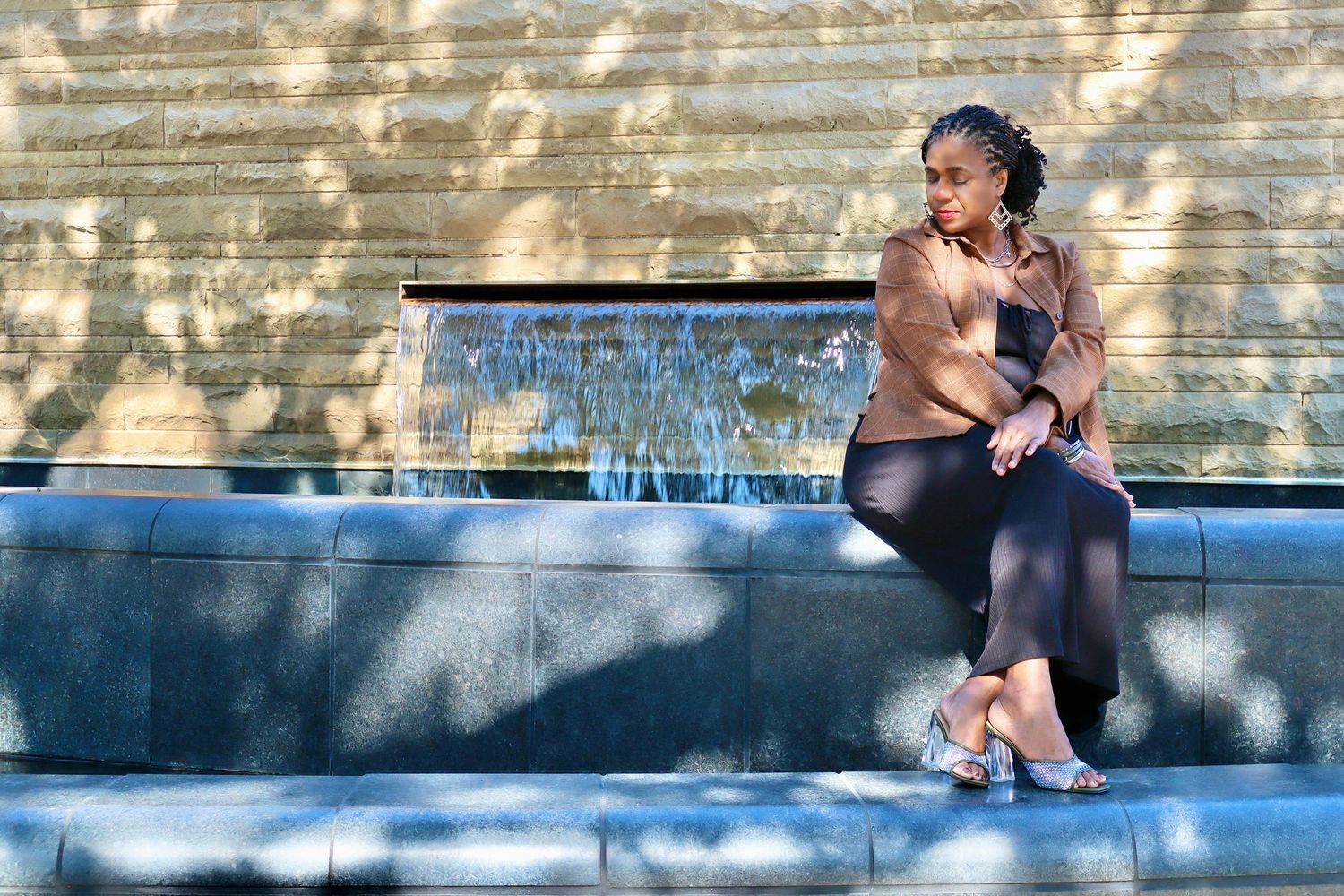 A woman sits thoughtfully on stone steps near a fountain, wearing a brown top and black pants in outdoor sunlight.