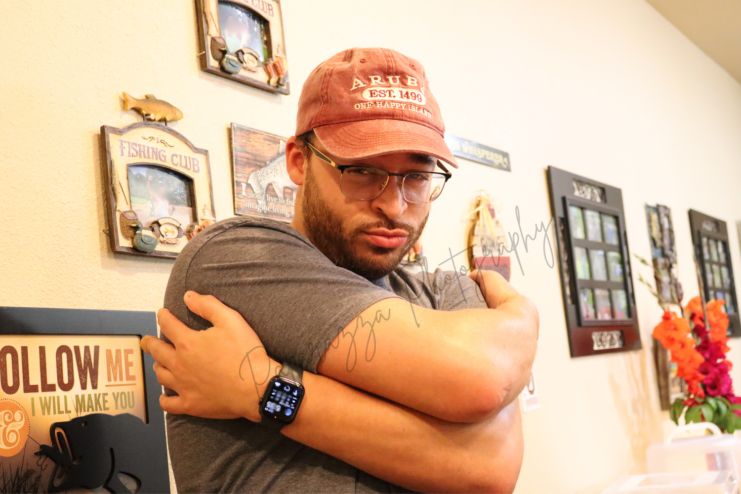 Bearded man in a red baseball cap, glasses and gray t-shirt crosses his arms over his chest in a room with framed wall decor.