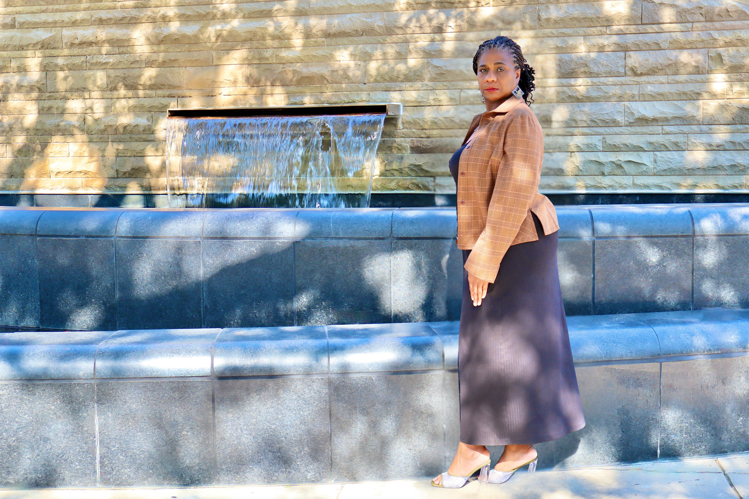 A woman in a brown blazer and dark skirt stands near a fountain in sunlight.