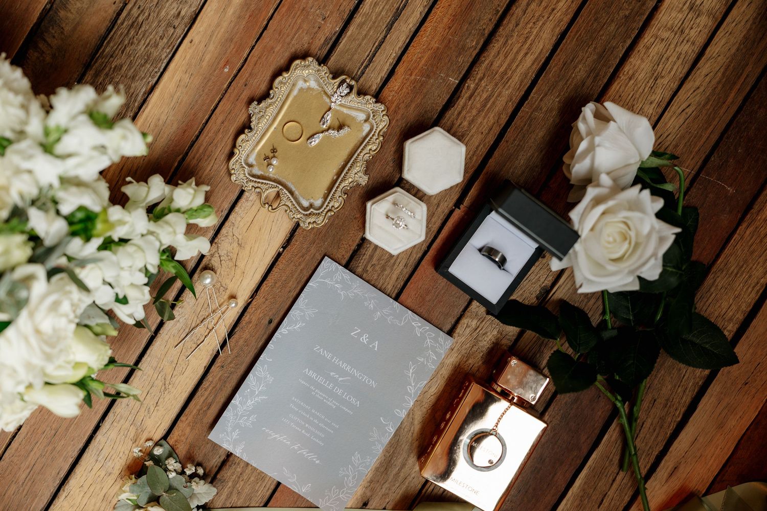 Elegant wedding flat lay with floral bouquet, rings, perfume, and gray invitation on rustic wood.