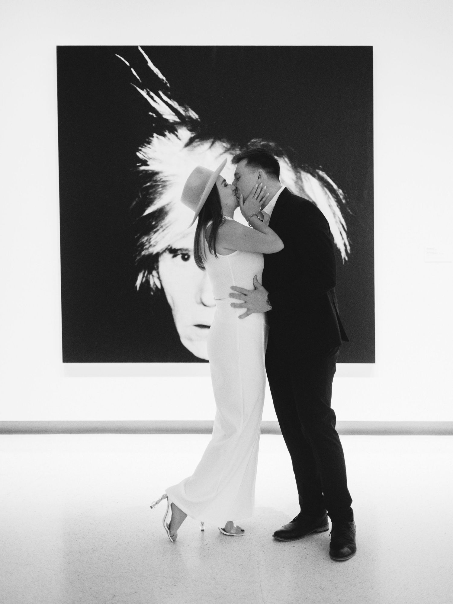 Couple kissing in white gallery before large Andy Warhol self-portrait painting, black and white photo.