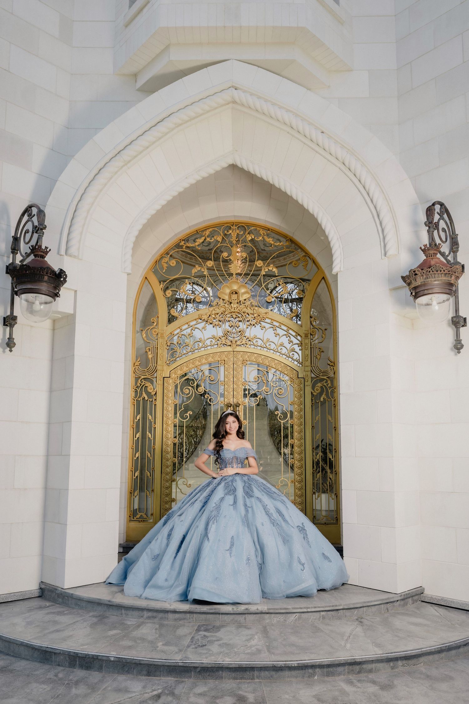 Young woman in a blue ball gown stands before ornate golden gates of a white castle-like building.