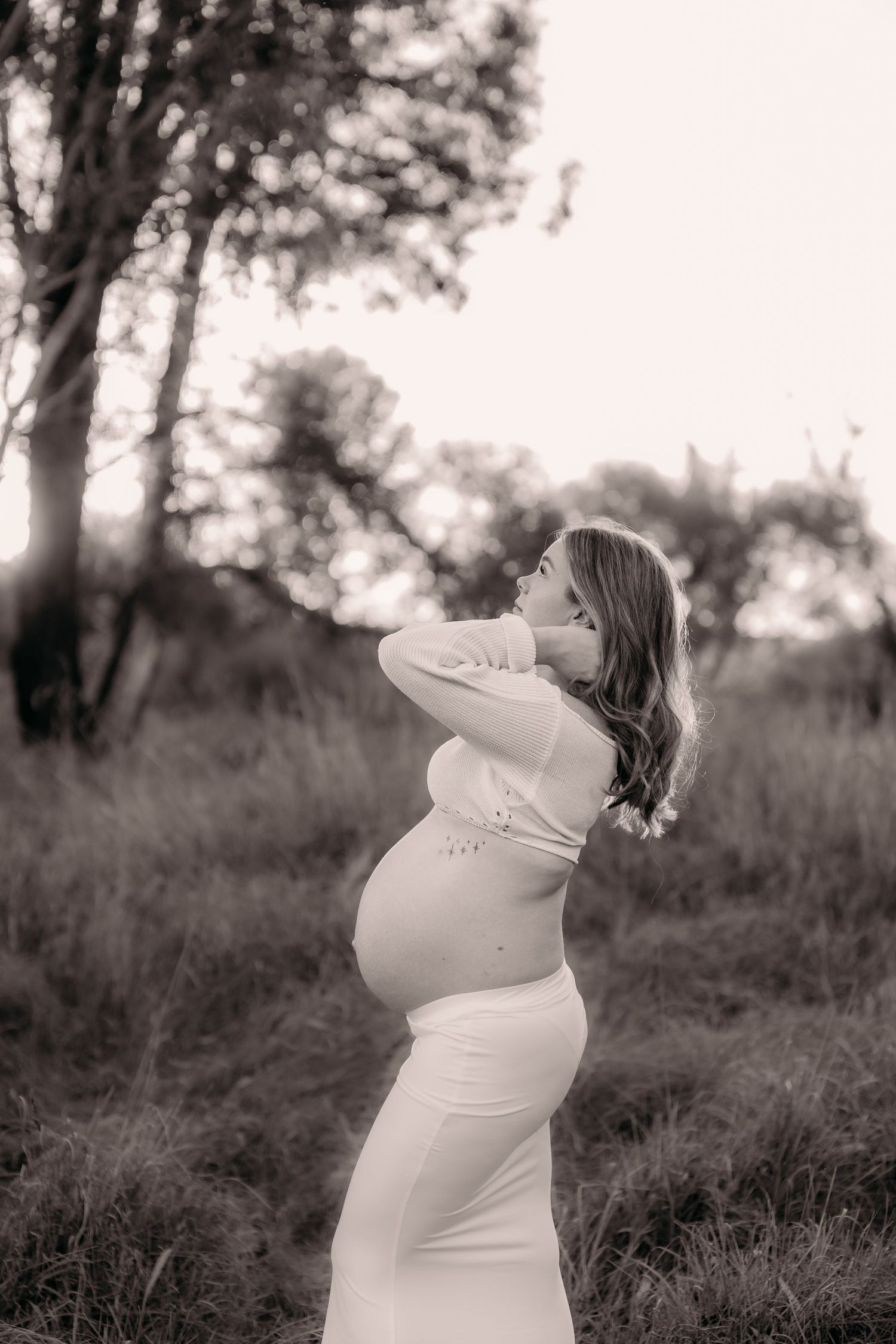 Black and white maternity photo of pregnant woman in white dress posing outdoors in nature at sunset.