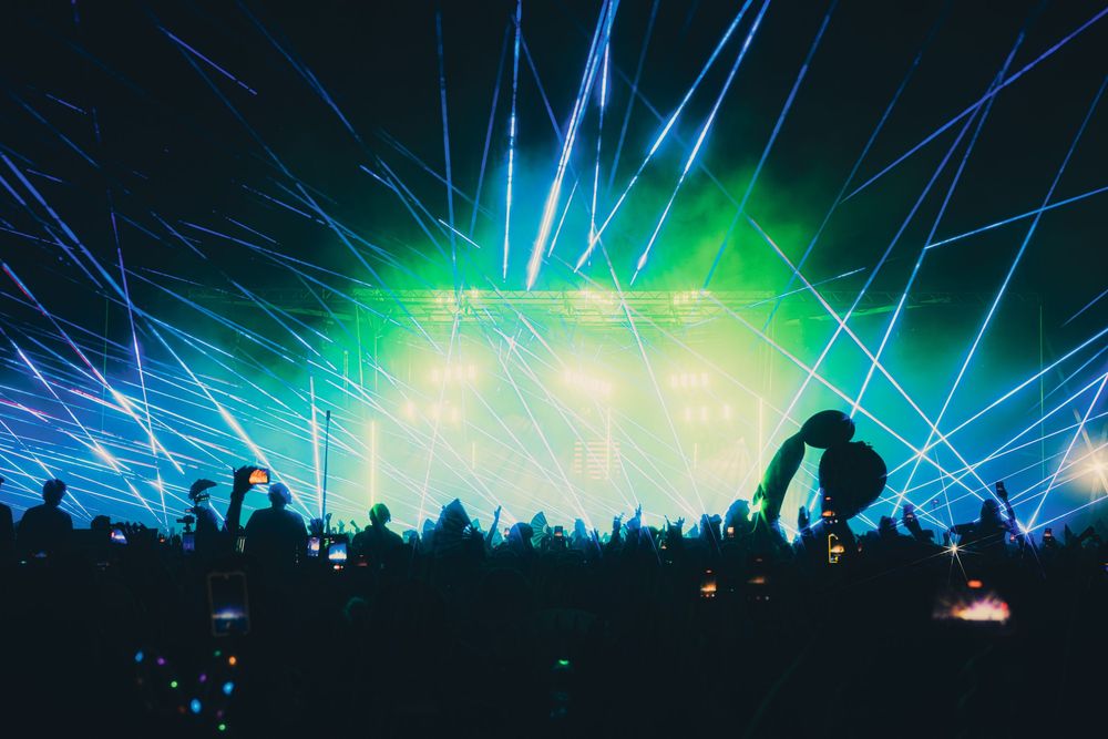A DJ performs on stage at an electronic music festival with vibrant green and blue laser lights illuminating the crowd.