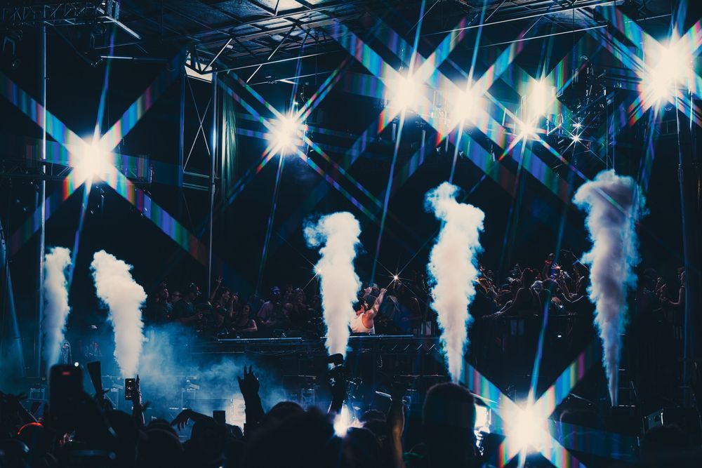 Concert stage with dramatic white smoke cannons, bright star-burst lights, and crowd silhouettes at a live music event.