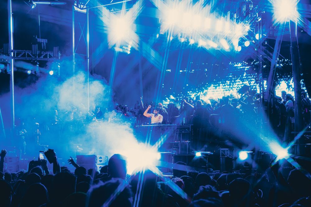 DJ performing on stage with dramatic blue laser lights and fog at a packed music festival concert.