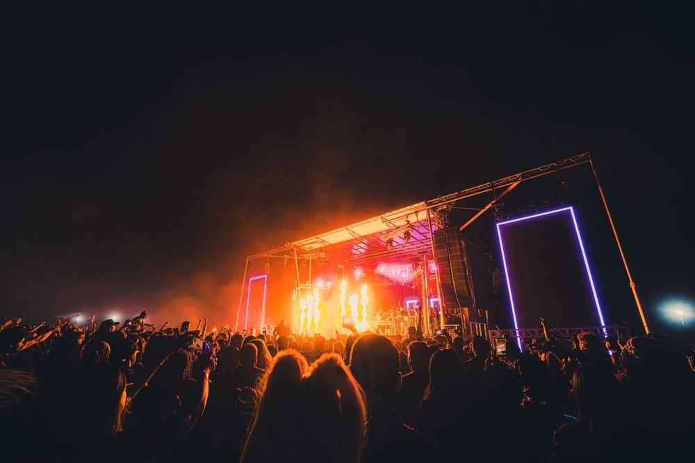 Crowd at outdoor night concert with vibrant red and purple stage lights illuminating the dark sky.