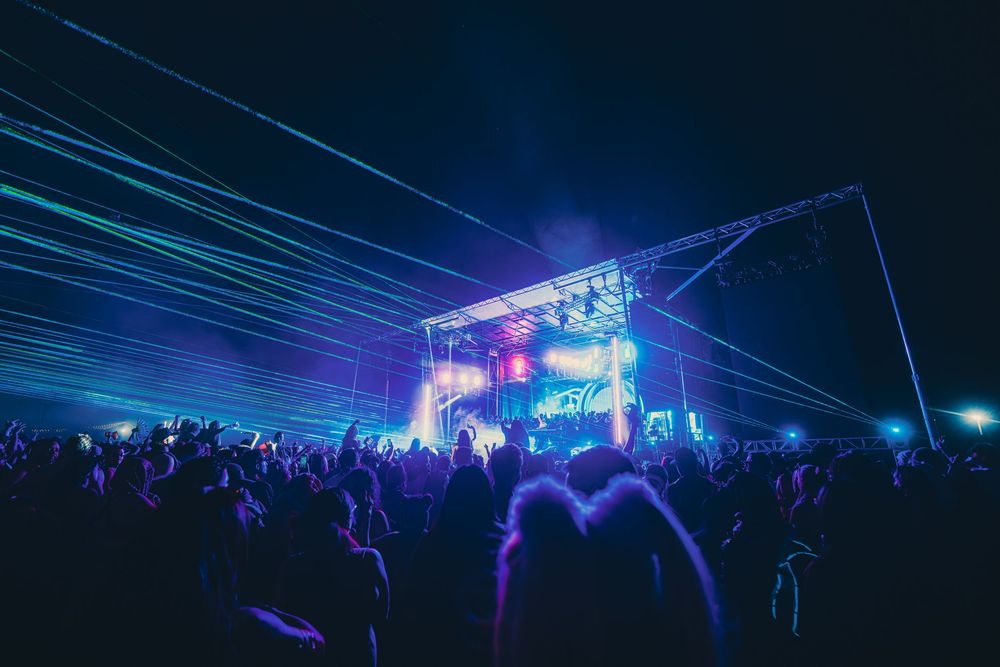 Night music festival crowd bathed in blue and purple laser lights with illuminated stage in background.