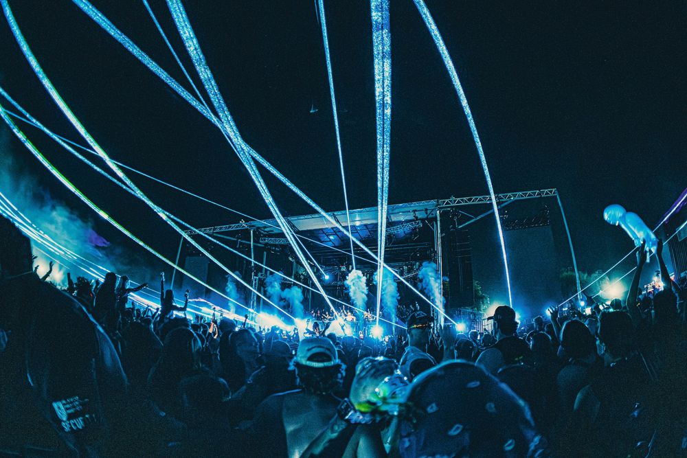 Crowd at an outdoor music festival at night with blue laser light show beams illuminating the sky.