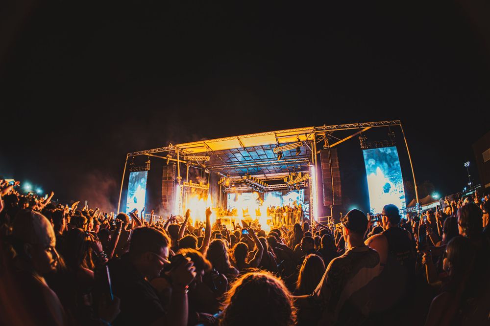 Large outdoor music festival crowd cheering in front of a brightly lit stage at night.