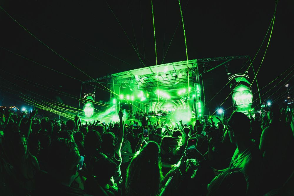 Massive outdoor music festival crowd bathed in green laser lights at night with illuminated stage.