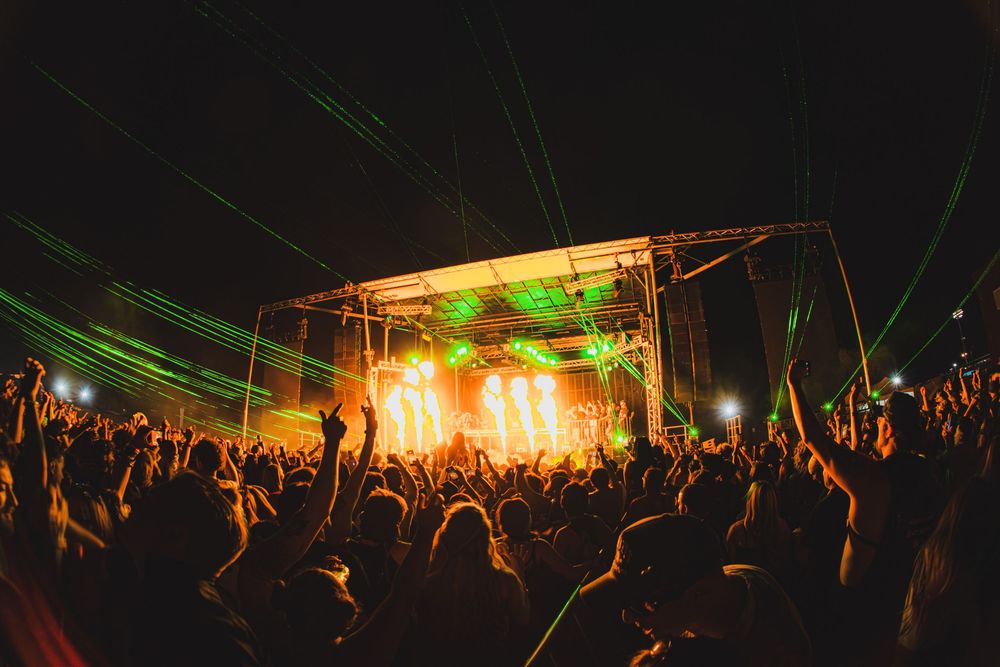 Vibrant green laser lights beam over a large crowd at an outdoor nighttime music festival with glowing stage.