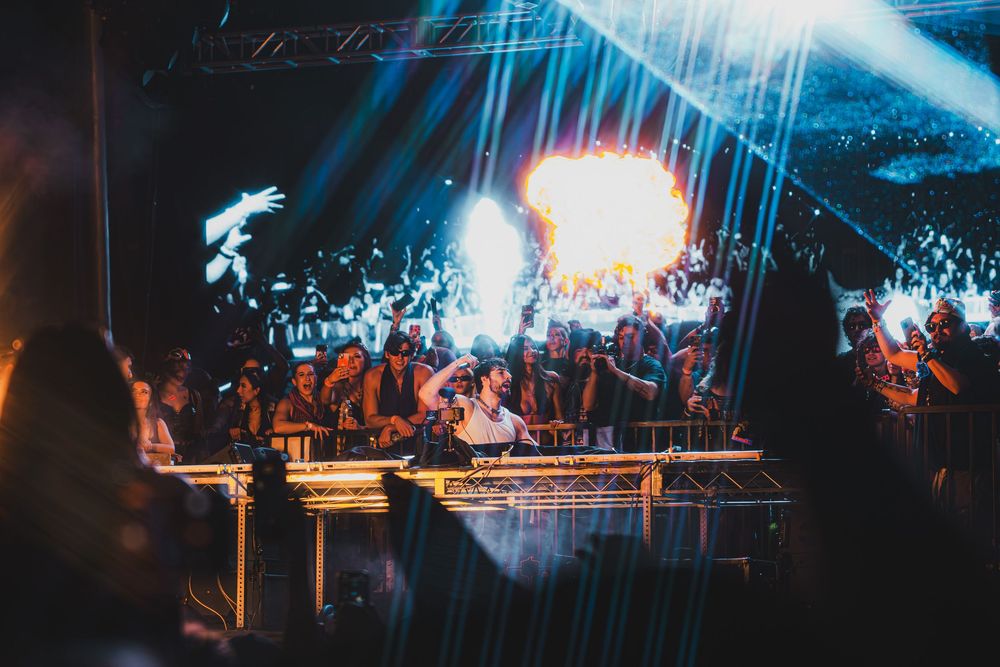 DJ performing on stage with dramatic fire effects and light beams at a crowded nightclub event.