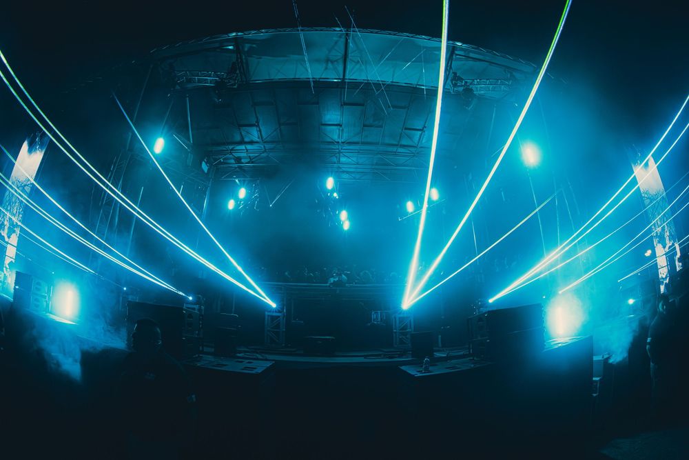 Blue laser light show illuminating a dark nightclub venue with foggy atmosphere and crowd silhouettes.