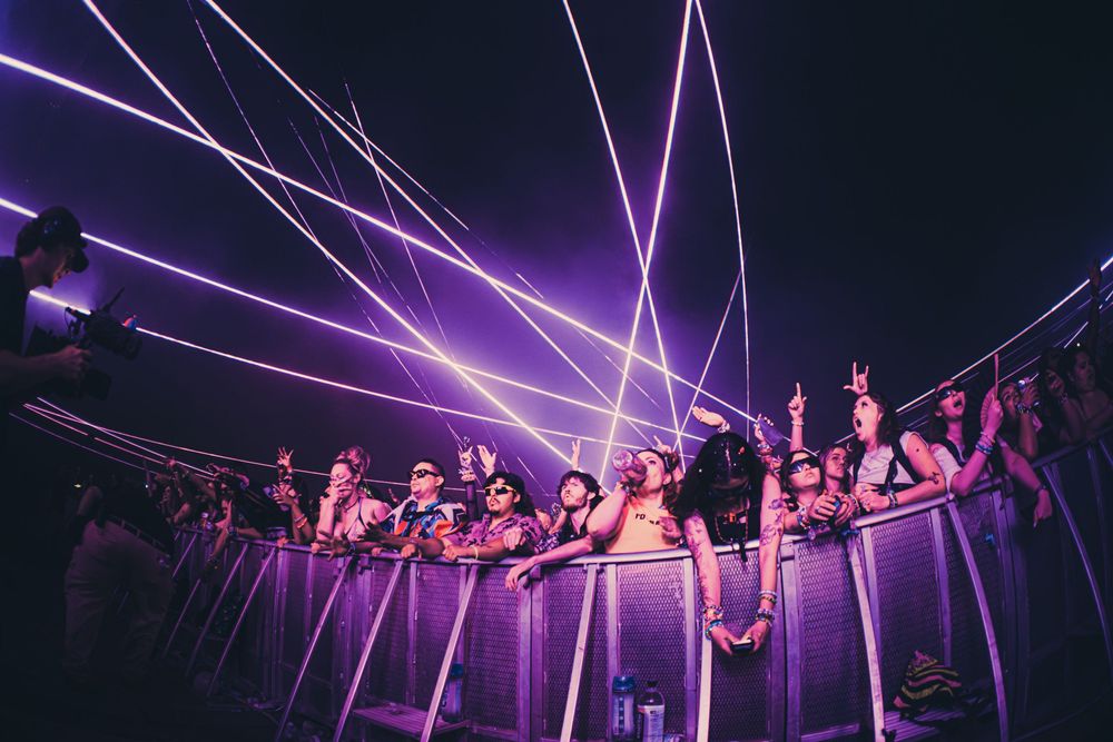 Concert crowd under purple laser lights at a music festival, fans cheering behind metal barriers at night.