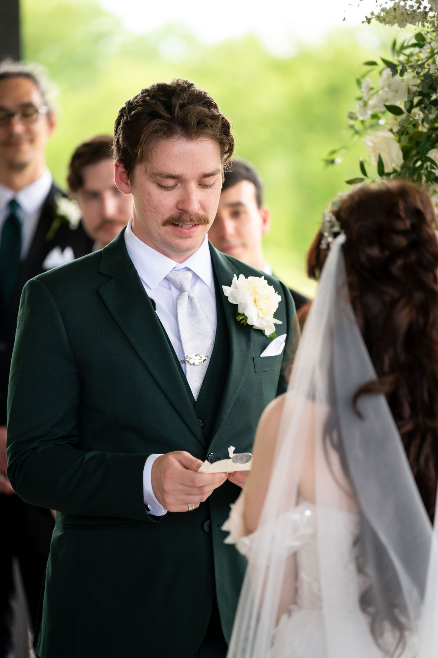 Groom in dark green suit reading vows to bride in white veil during outdoor wedding ceremony.
