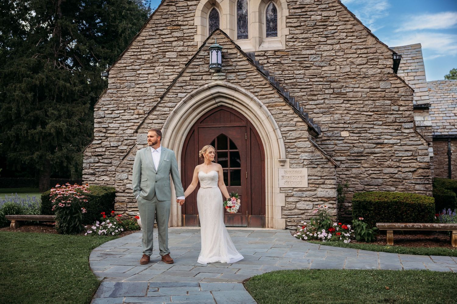 Bride and groom posing in front of a historic stone church with arched wooden doors and rose gardens.
