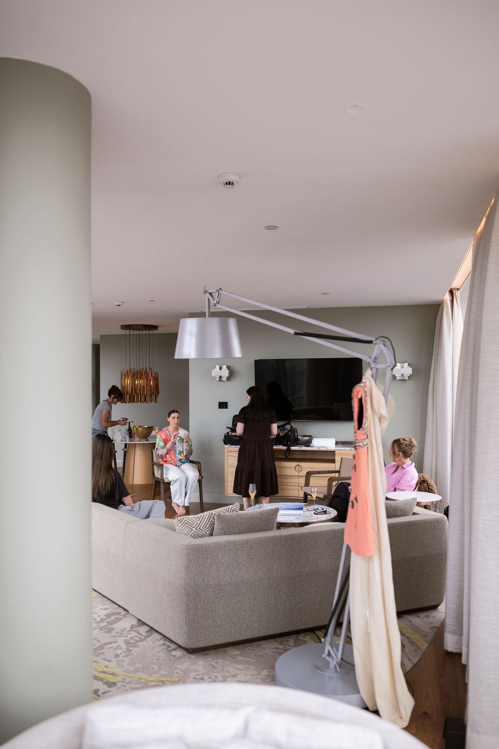 Modern hotel suite lounge with guests relaxing on neutral sofas, flat screen TV, and elegant pendant lighting.