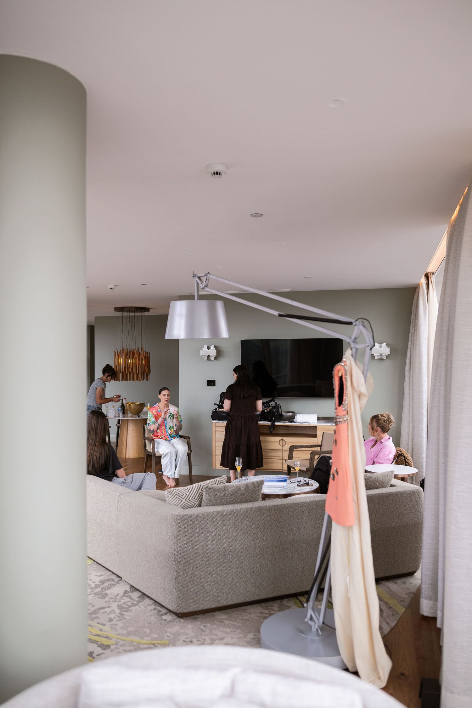 Modern hotel suite lounge with guests relaxing on neutral sofas, flat screen TV, and elegant pendant lighting.