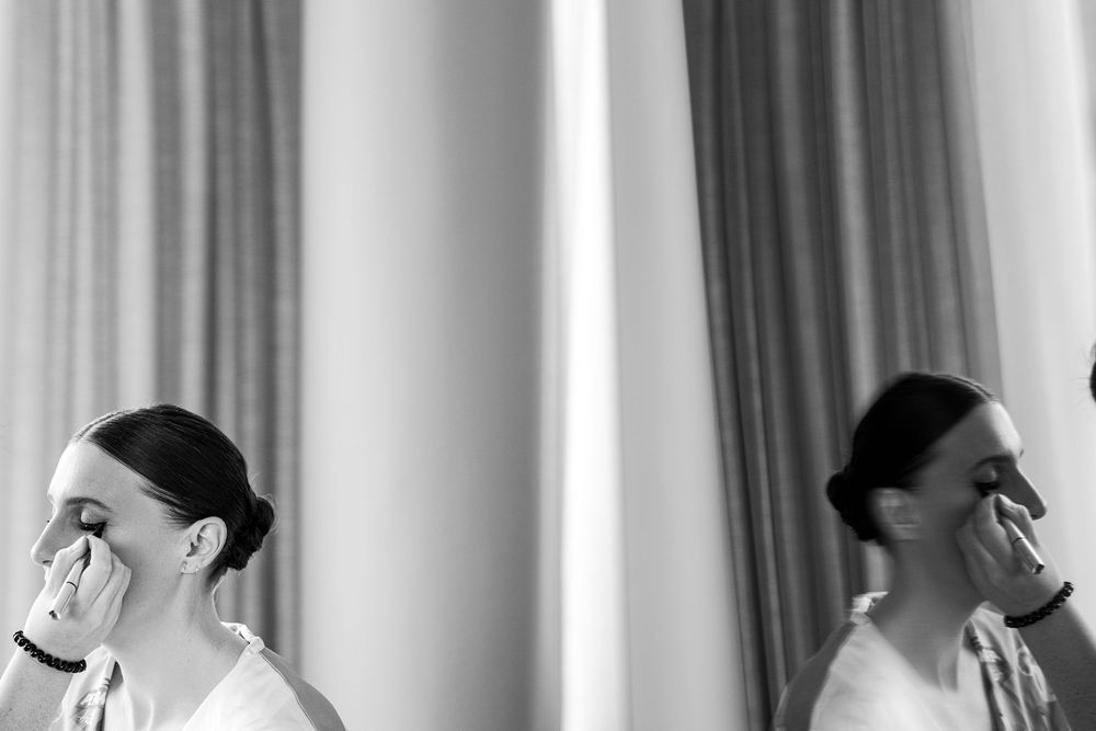 Black and white photo of a bride applying makeup, reflected in a mirror, capturing elegant wedding preparation moments.