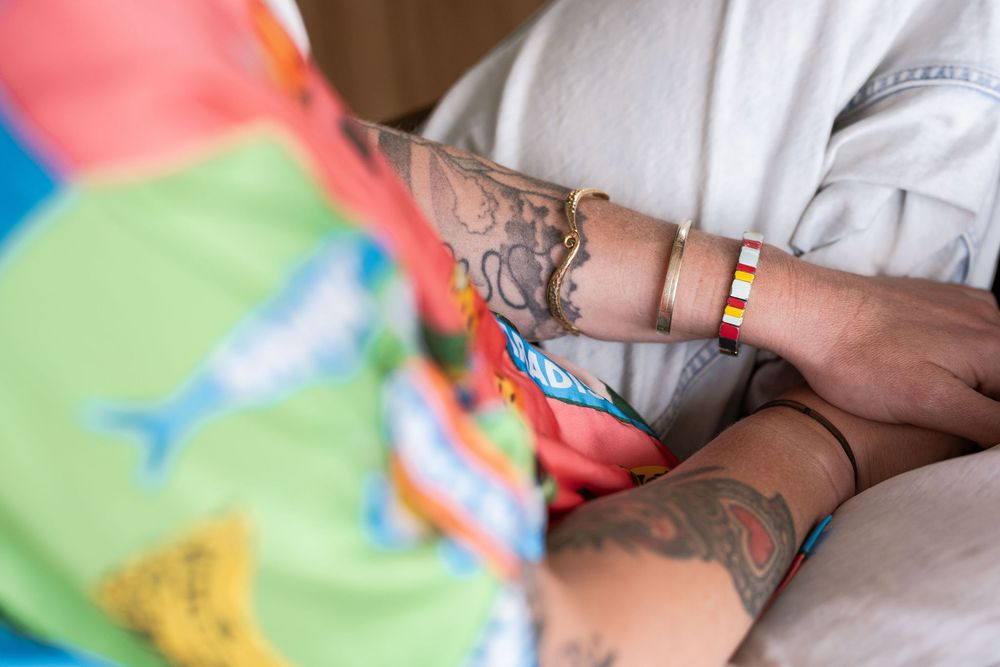 Close-up of tattooed arms with colorful bracelets, wearing a white shirt, next to a vibrant patterned fabric.