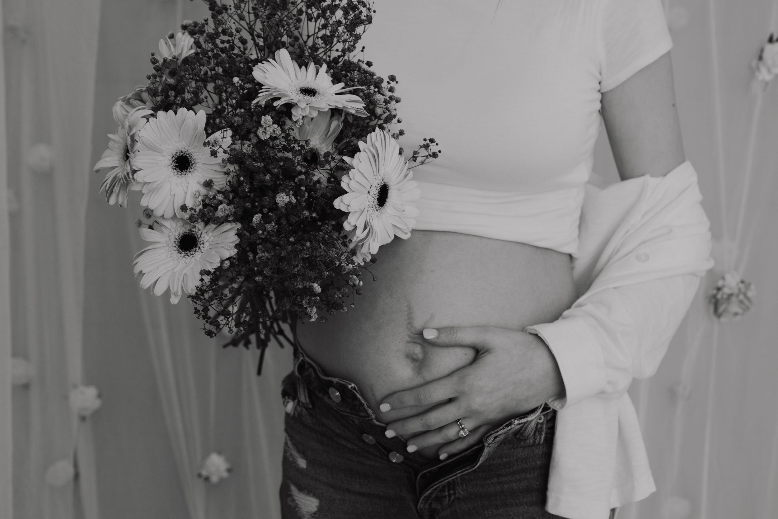 Black and white photo of pregnant woman holding flowers, cradling baby bump in jeans and white crop top.