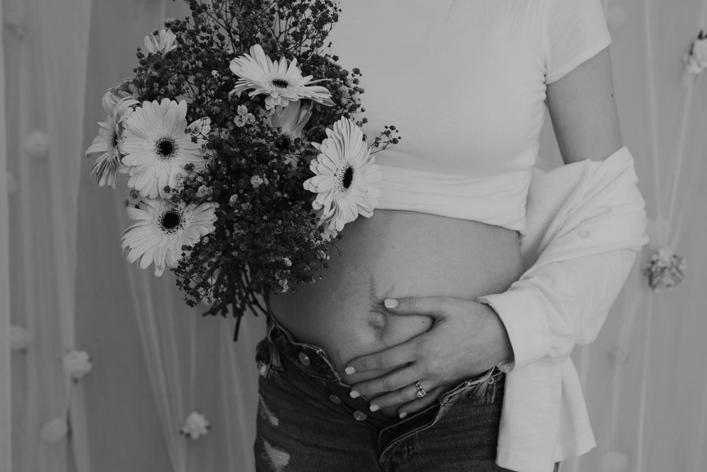 Black and white photo of pregnant woman holding flowers, cradling baby bump in jeans and white crop top.