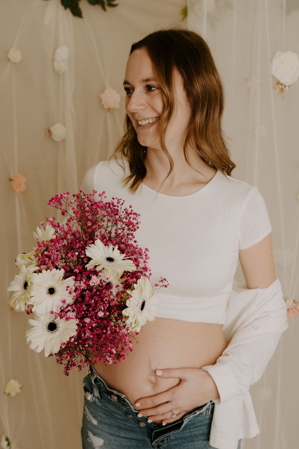 Smiling pregnant woman in white crop top holding pink and white flower bouquet while cradling baby bump in floral studio.
