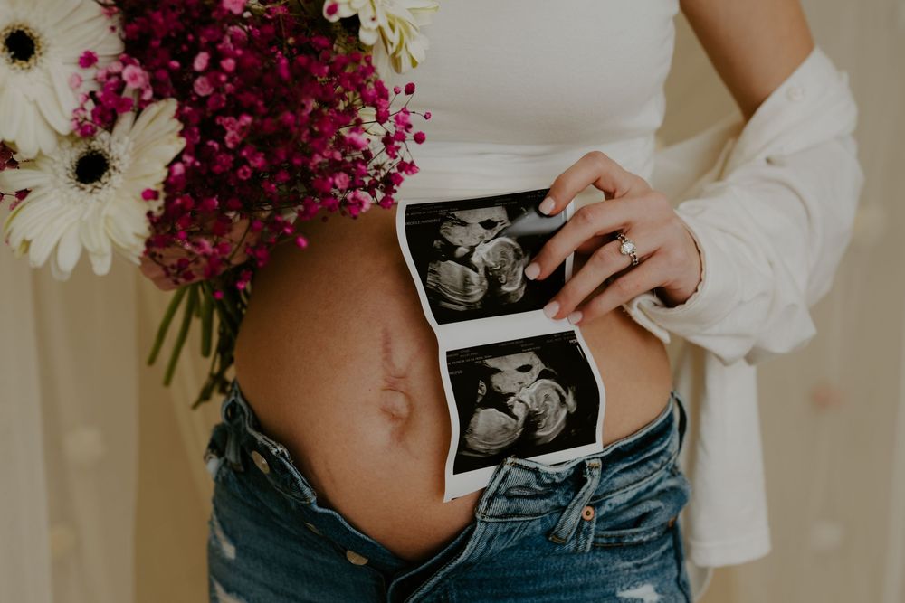 Pregnant woman holding ultrasound photos and flowers, showing bare baby bump in white top and ripped jeans.