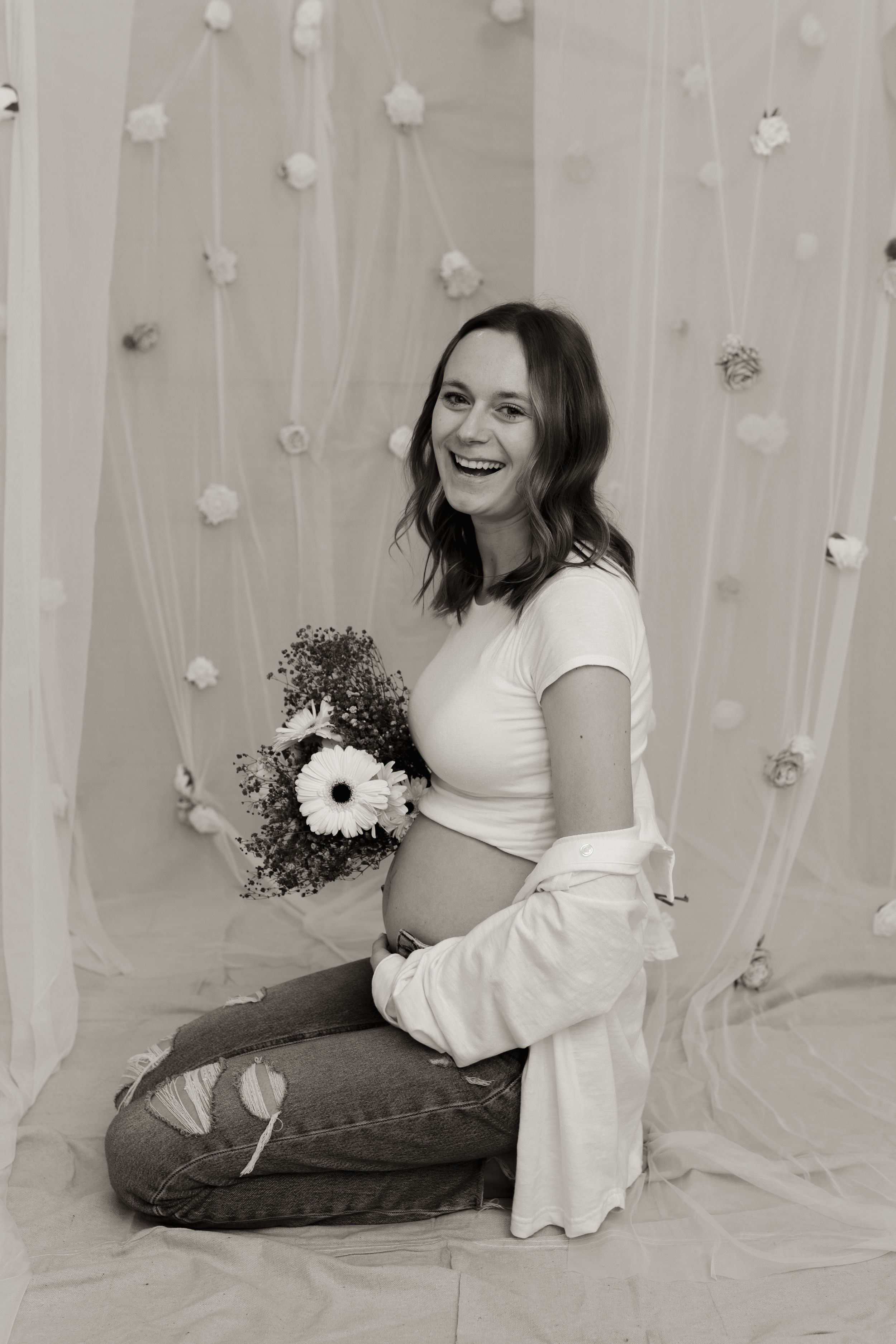 Black and white photo of a smiling pregnant woman sitting on floor holding flowers, wearing jeans and a white top.