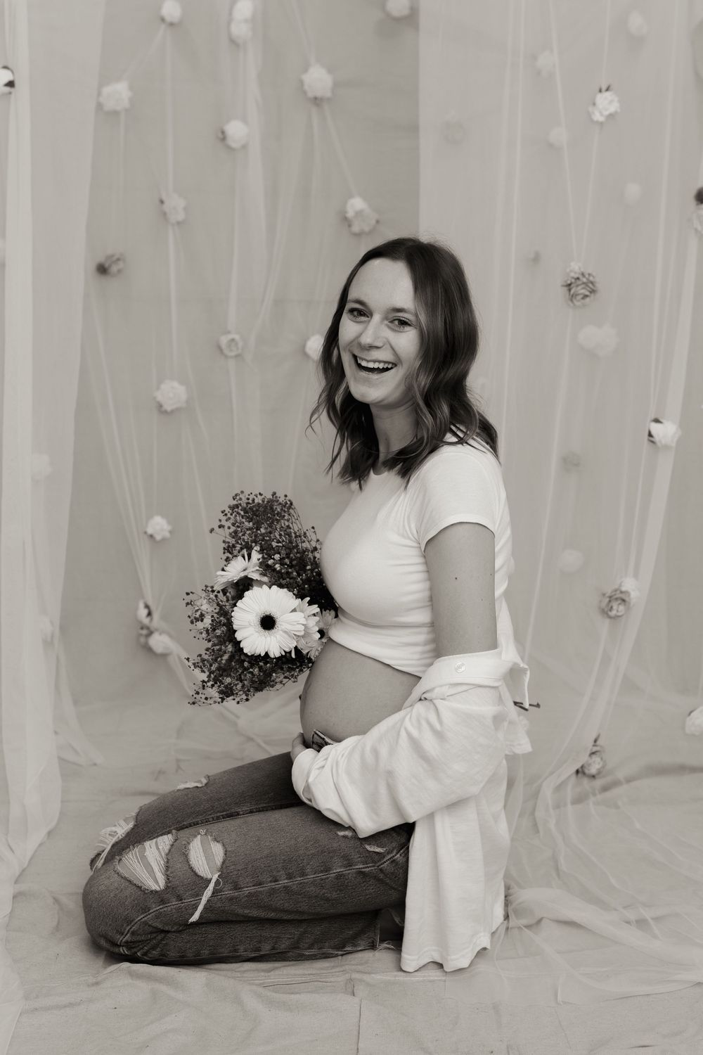 Black and white photo of a smiling pregnant woman sitting on floor holding flowers, wearing jeans and a white top.