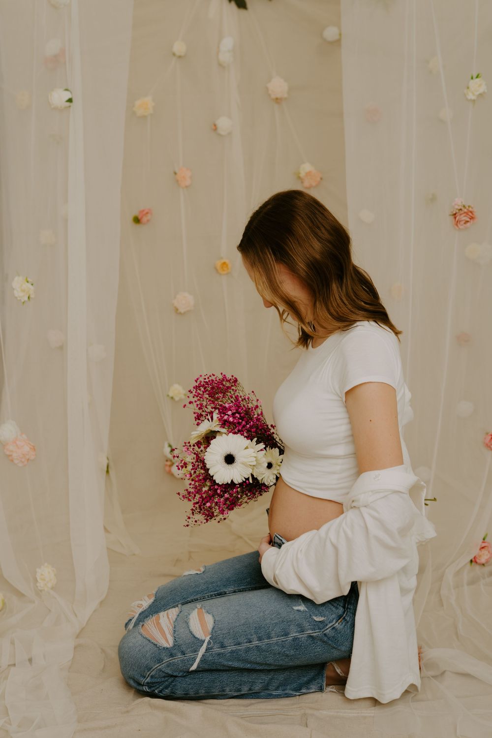 Pregnant woman kneeling in white crop top and jeans, holding pink and white flowers against a floral backdrop.